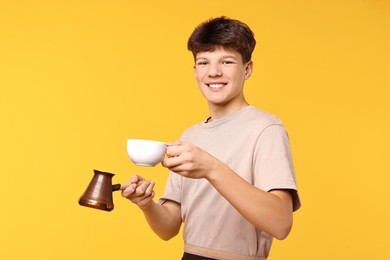 Photo of Teenage boy with cezve pot and cup working as barista on orange background