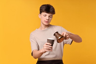 Photo of Teenage boy with cezve pot and paper cup working as barista on orange background