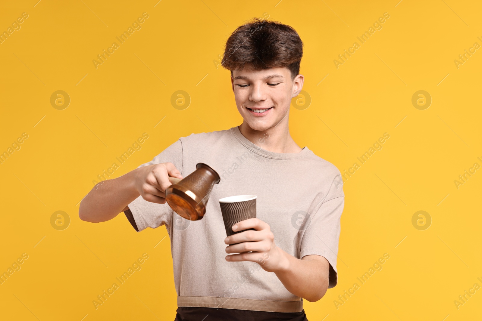 Photo of Teenage boy with cezve pot and paper cup working as barista on orange background