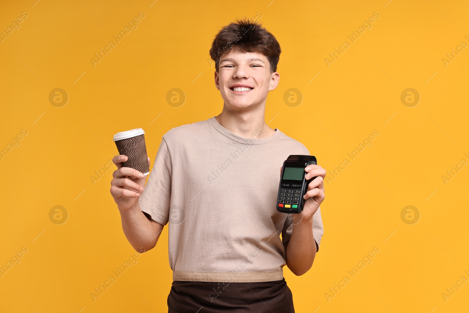 Photo of Teenage boy with paper cup and payment terminal working as barista on orange background