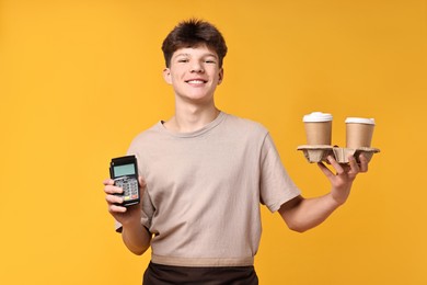 Photo of Teenage boy with paper cups and payment terminal working as barista on orange background