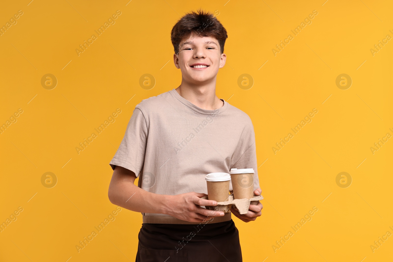 Photo of Teenage boy with paper cups working as barista on orange background