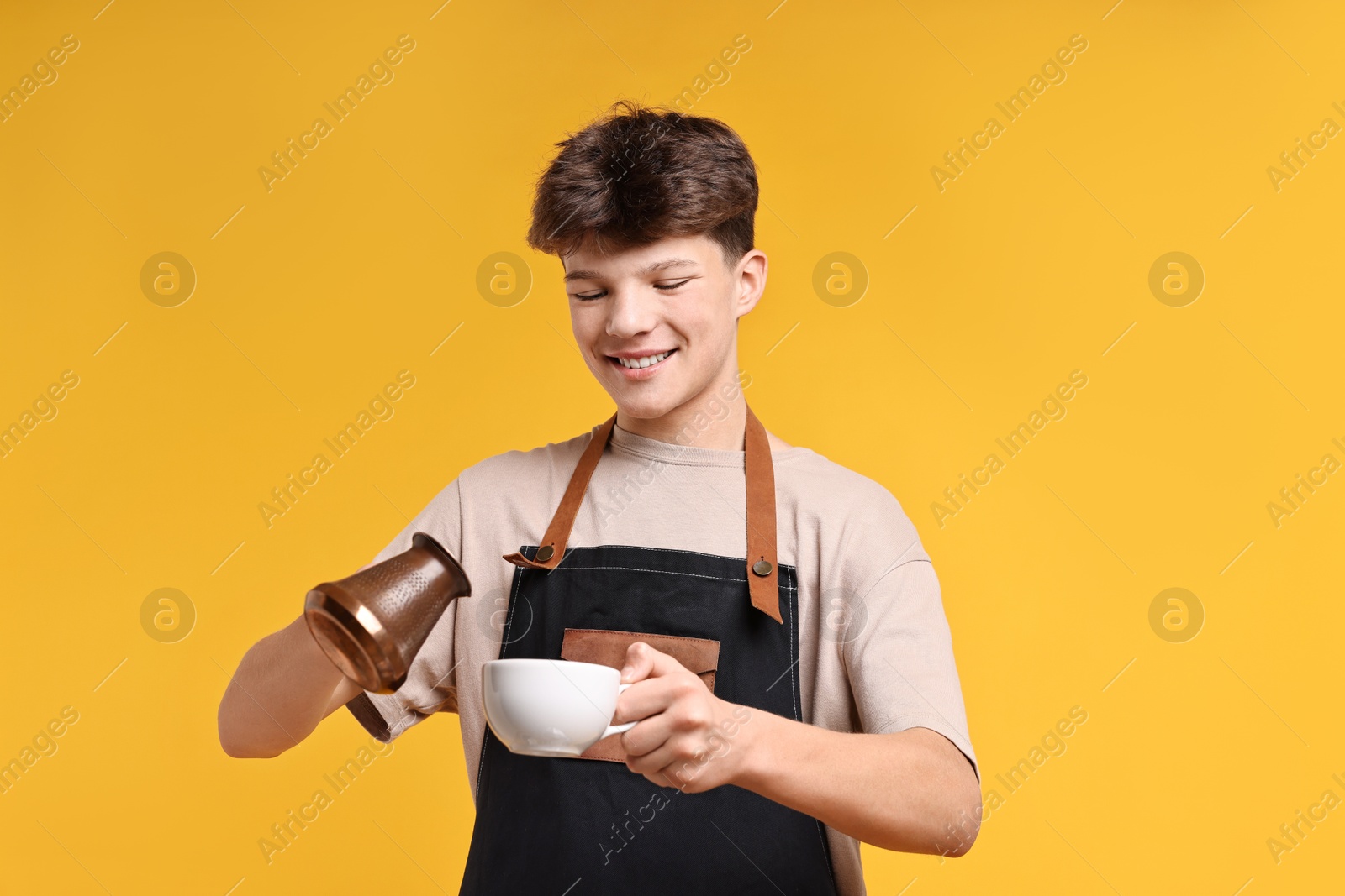 Photo of Teenage boy with cezve pot and cup working as barista on orange background