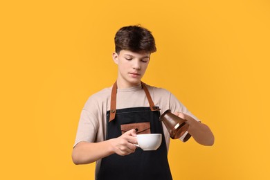 Photo of Teenage boy with cezve pot and cup working as barista on orange background