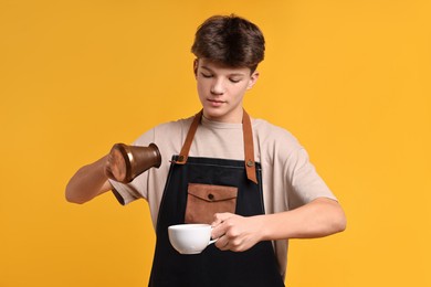 Photo of Teenage boy with cezve pot and cup working as barista on orange background