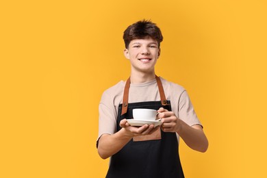 Photo of Teenage boy with cup of coffee working as barista on orange background