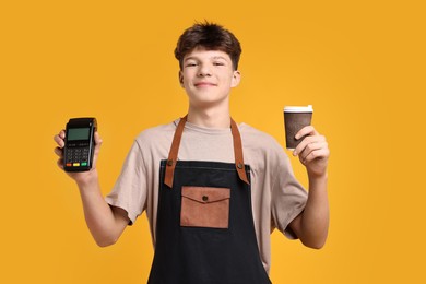 Photo of Teenage boy with paper cup and payment terminal working as barista on orange background