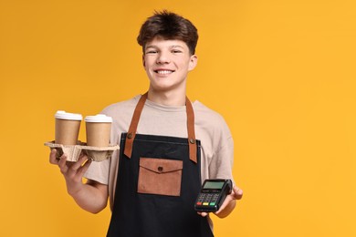 Photo of Teenage boy with paper cups and payment terminal working as barista on orange background