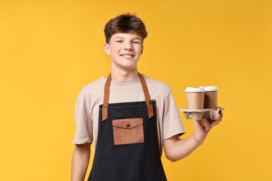 Photo of Teenage boy with paper cups working as barista on orange background