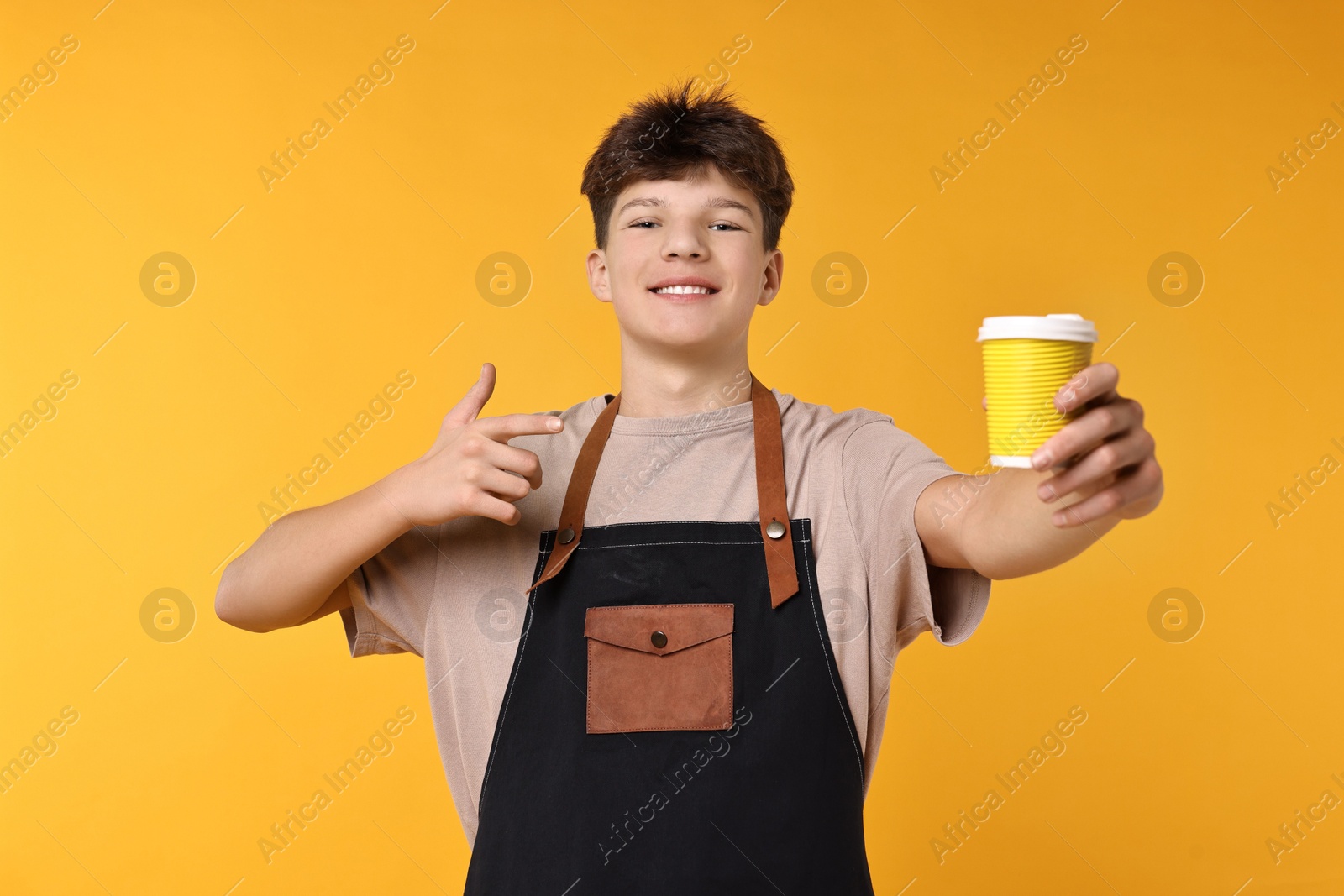 Photo of Teenage boy with cup of coffee working as barista on orange background