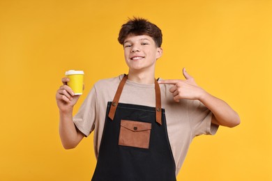 Photo of Teenage boy with cup of coffee working as barista on orange background