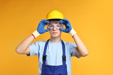 Photo of Teenage boy in hardhat and protective mask working as builder on orange background