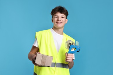 Photo of Teenage boy with tape gun dispenser and box in safety vest working as warehouse packer on blue background