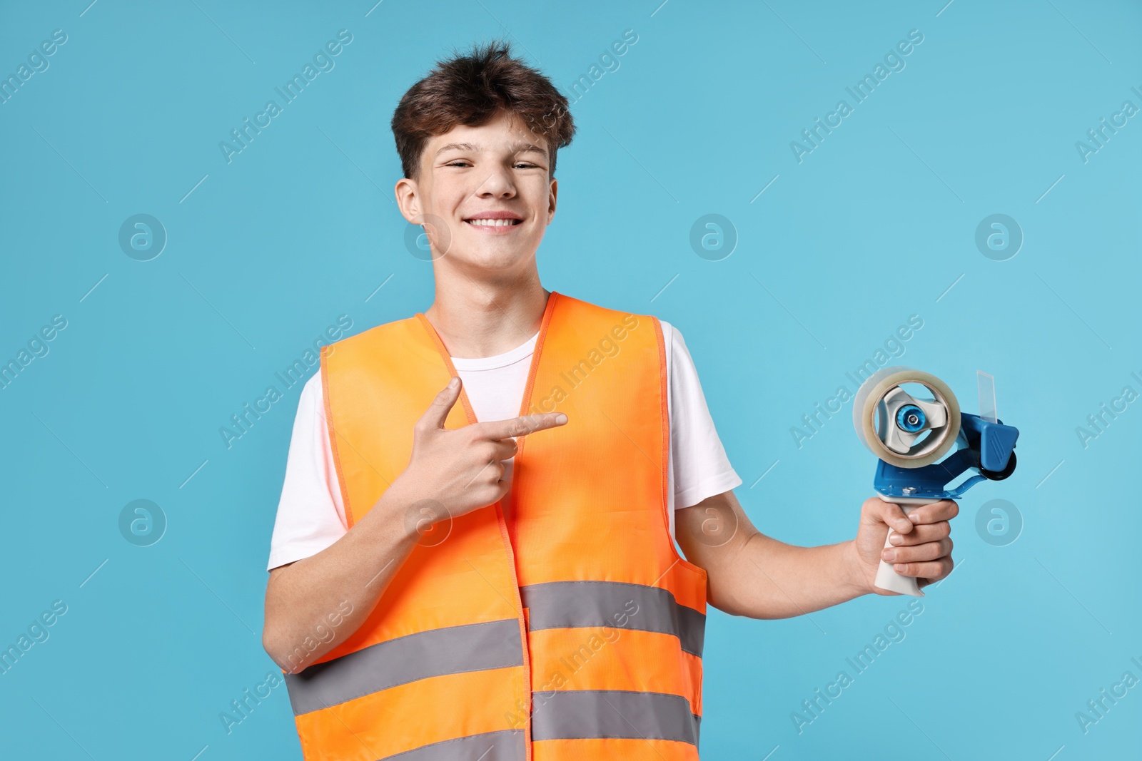 Photo of Teenage boy with tape gun dispenser in safety vest working as warehouse packer on blue background