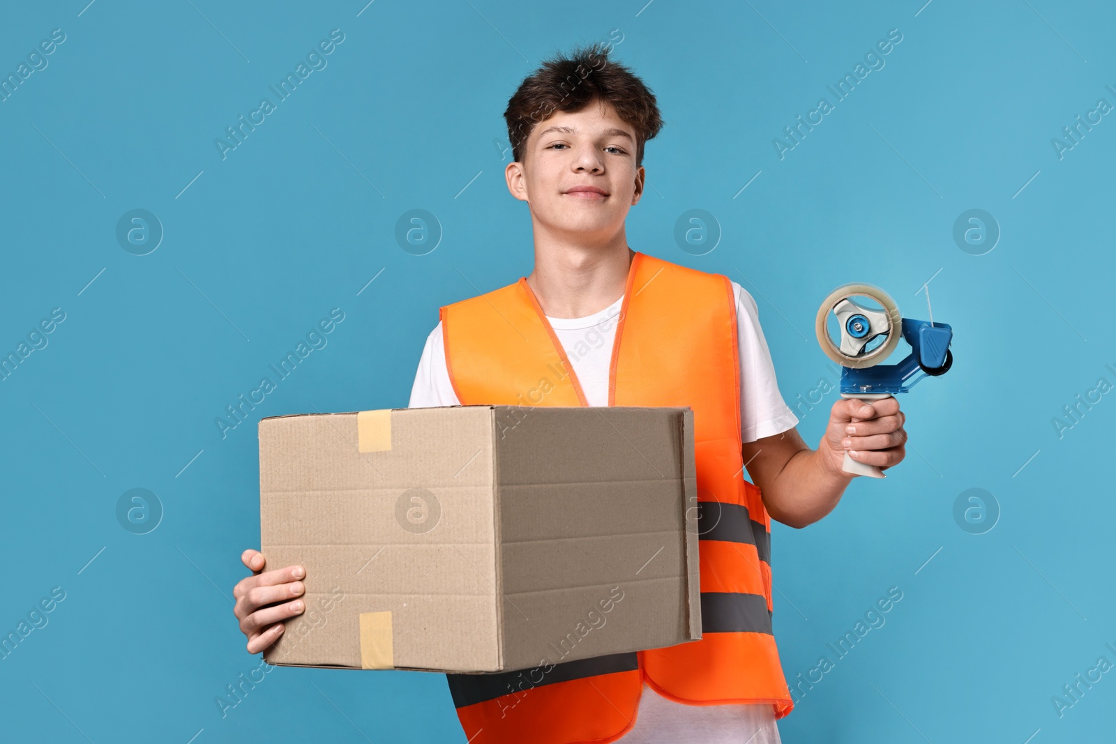 Photo of Teenage boy with tape gun dispenser and box in safety vest working as warehouse packer on blue background