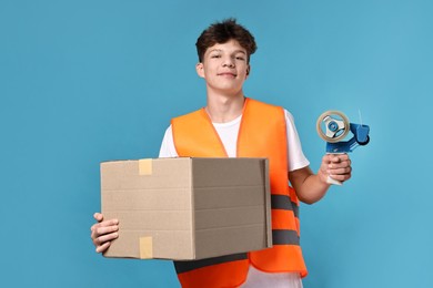 Photo of Teenage boy with tape gun dispenser and box in safety vest working as warehouse packer on blue background