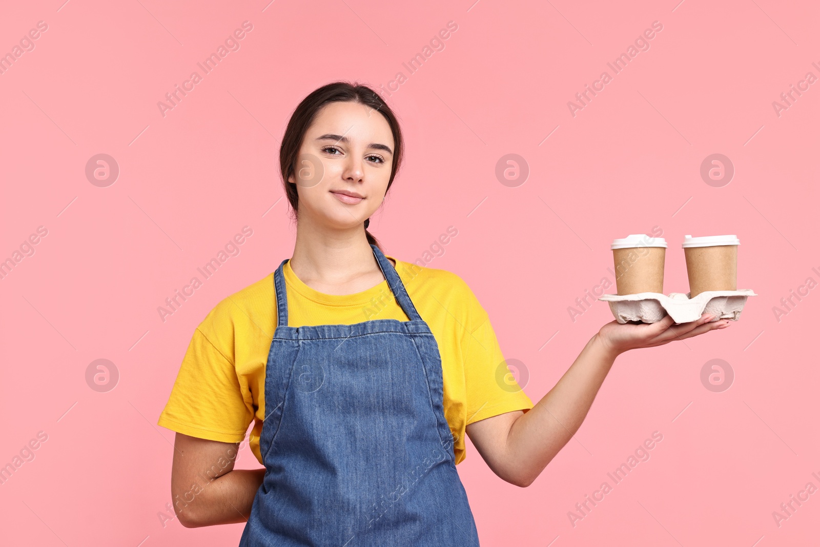 Photo of Girl in apron with takeaway paper cups of coffee on pink background. Work for teenagers