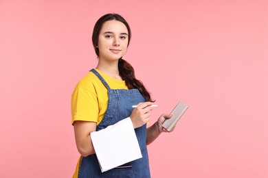 Photo of Girl in apron with pen and notebook on pink background. Work for teenagers