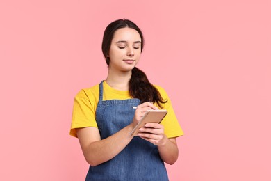 Photo of Girl in apron with pen and notebook on pink background. Work for teenagers