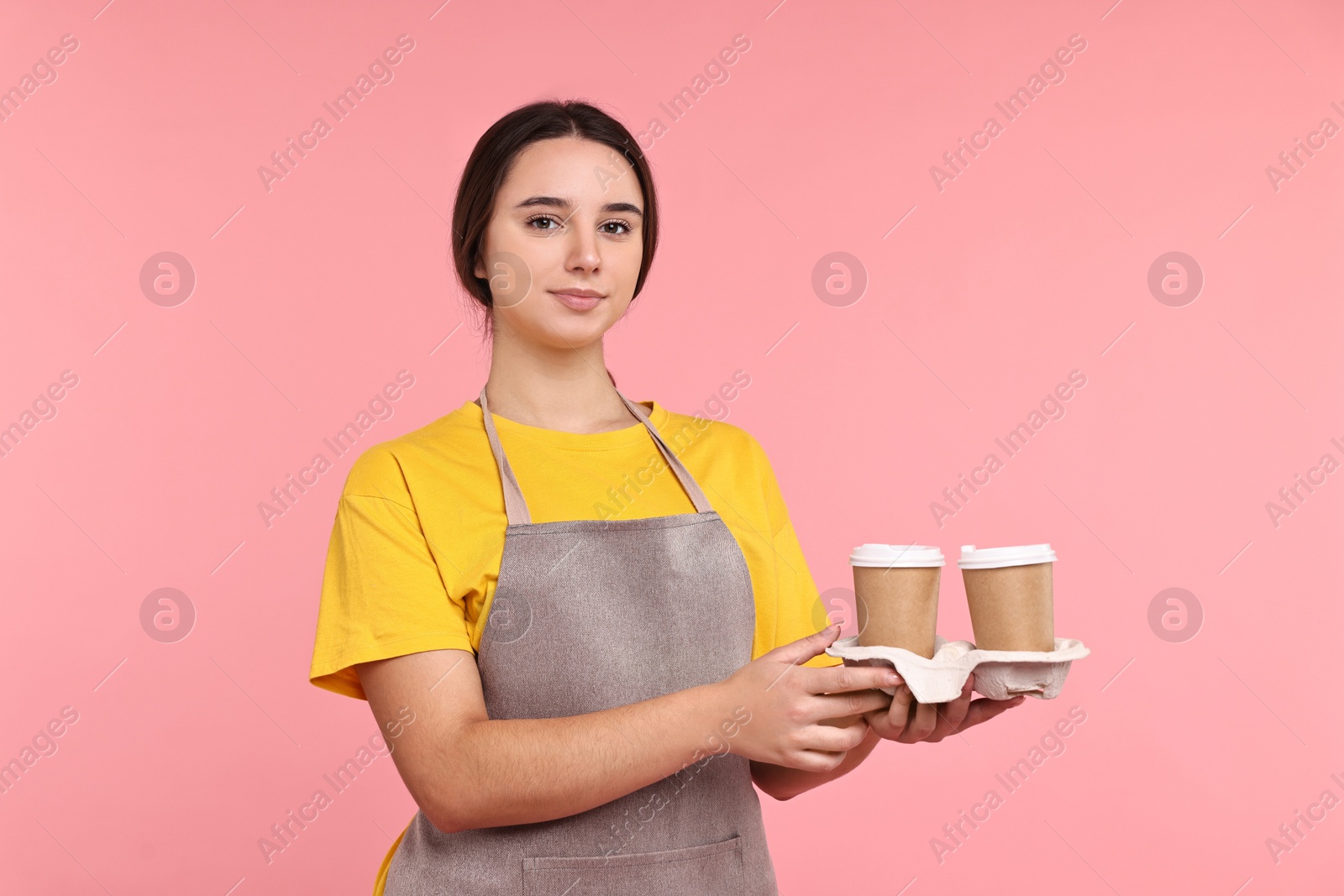 Photo of Girl in apron with takeaway paper cups of coffee on pink background. Work for teenagers