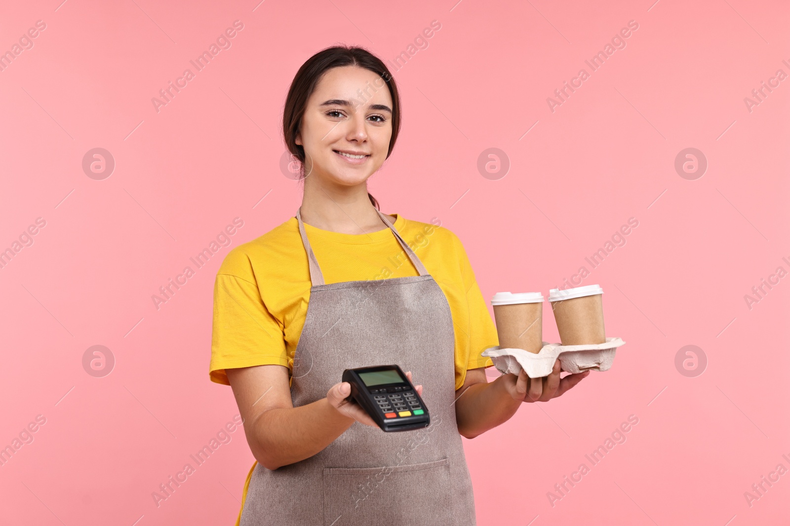 Photo of Girl in apron with paper cups of coffee and payment terminal on pink background. Work for teenagers