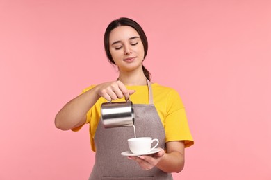 Photo of Girl in apron pouring milk from pitcher into cup of coffee on pink background. Work for teenagers