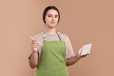 Photo of Girl in apron with pen and notebook on pale brown background. Work for teenagers