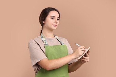 Photo of Girl in apron with pen and notebook on pale brown background. Work for teenagers