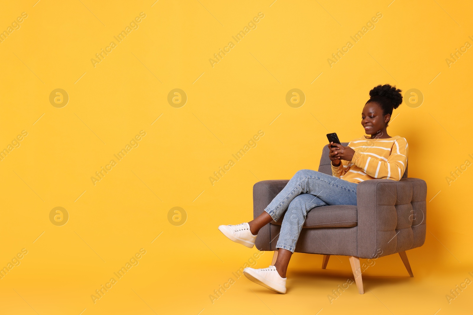 Photo of Smiling woman with smartphone sitting in armchair on yellow background. Space for text