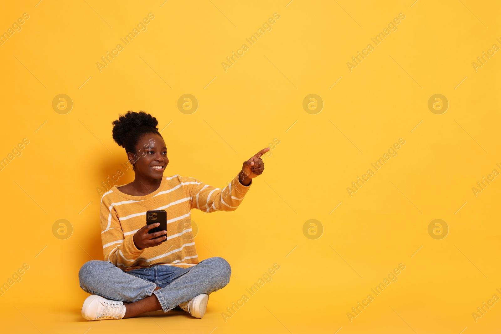 Photo of Cheerful woman with smartphone pointing at something on yellow background. Space for text