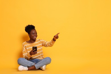 Photo of Cheerful woman with smartphone pointing at something on yellow background. Space for text