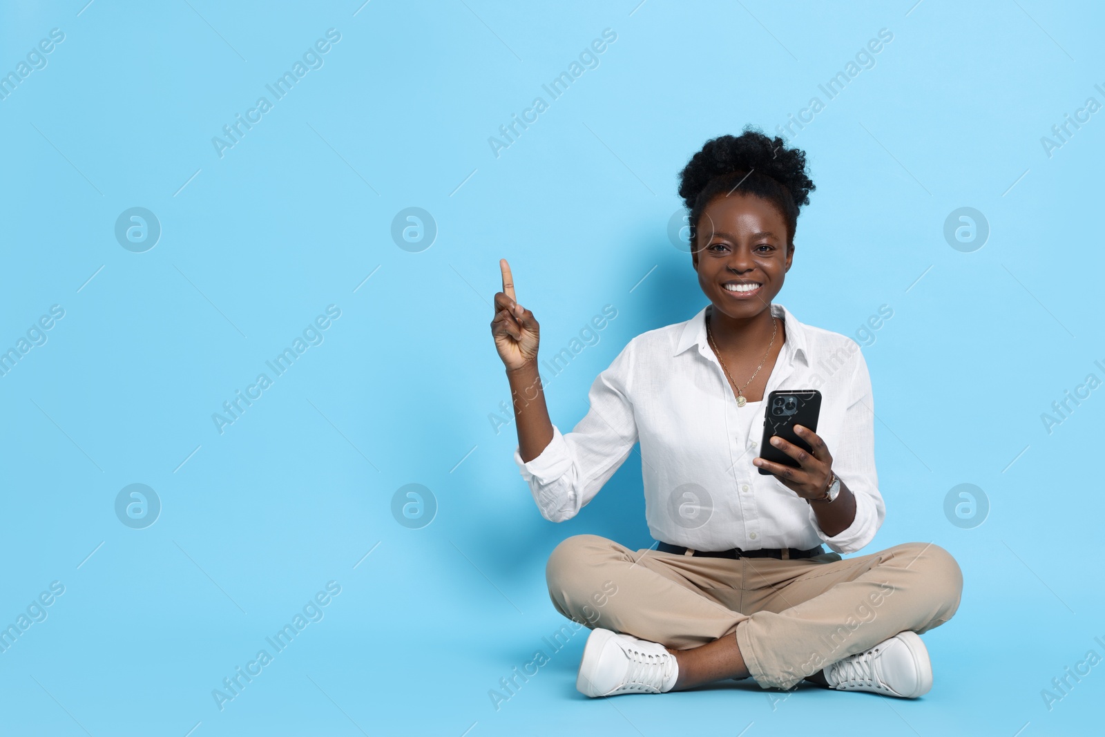 Photo of Cheerful woman with smartphone pointing at something on light blue background. Space for text