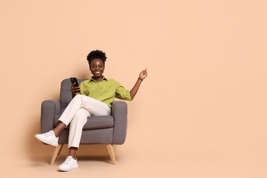 Cheerful woman with smartphone sitting in armchair and pointing at something on beige background. Space for text