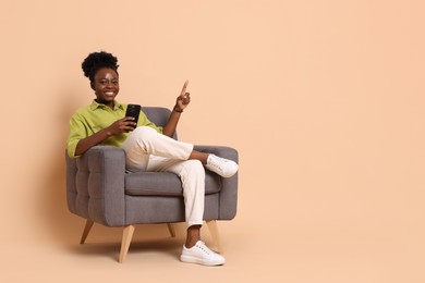 Cheerful woman with smartphone sitting in armchair and pointing at something on beige background. Space for text