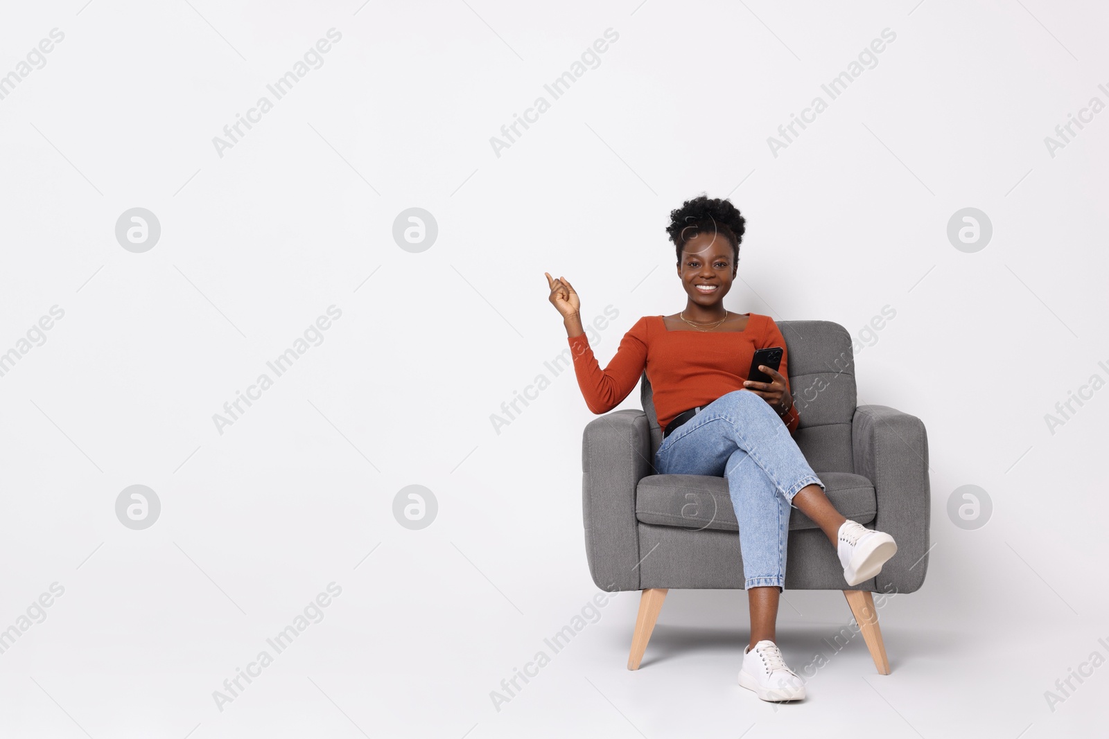 Photo of Cheerful woman with smartphone sitting in armchair and pointing at something on white background. Space for text