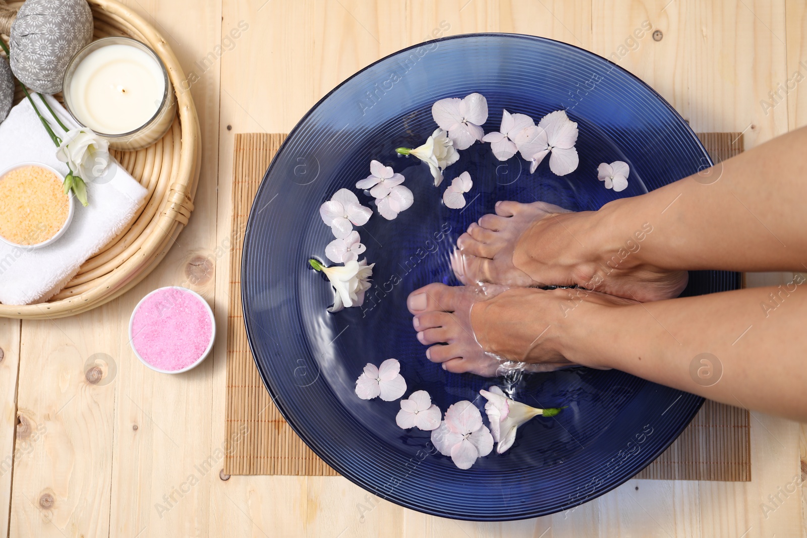 Photo of Woman soaking her feet in bowl with water and flowers on floor, top view. Spa treatment