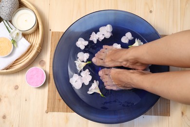 Photo of Woman soaking her feet in bowl with water and flowers on floor, top view. Spa treatment