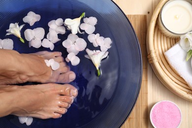 Photo of Woman soaking her feet in bowl with water and flowers on floor, top view. Spa treatment