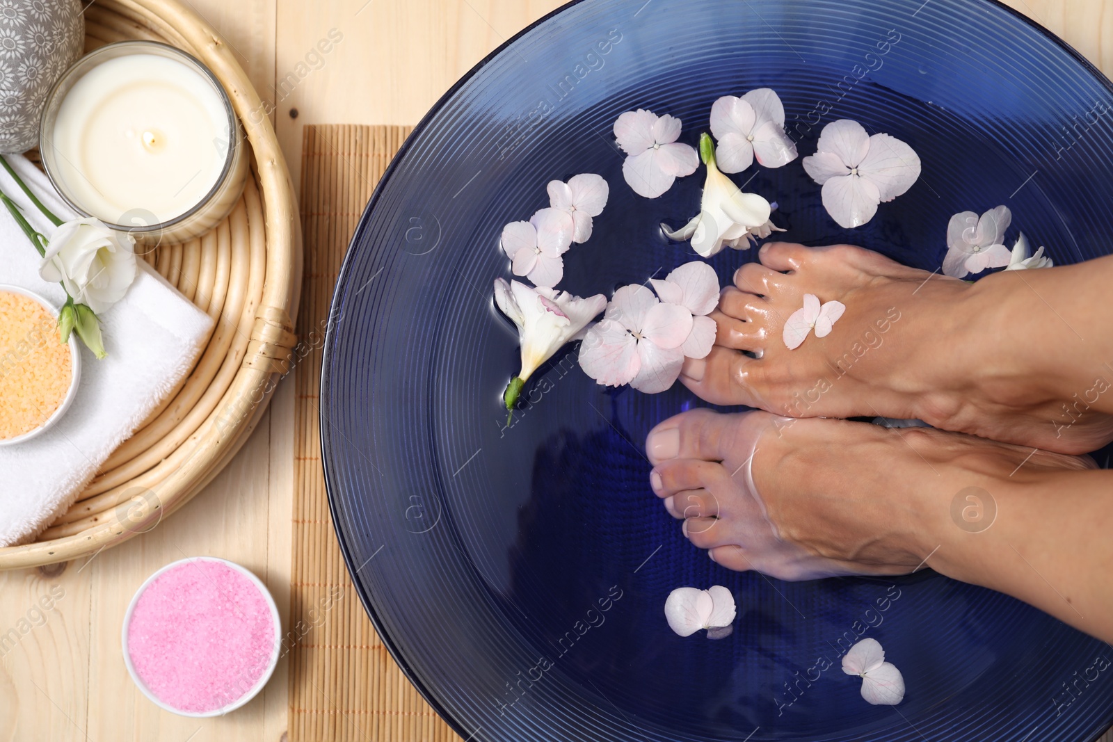 Photo of Woman soaking her feet in bowl with water and flowers on floor, top view. Spa treatment