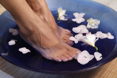 Photo of Woman soaking her feet in bowl with water and flowers on floor, closeup. Spa treatment