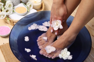 Photo of Woman soaking her feet in bowl with water and flowers on floor, closeup. Spa treatment