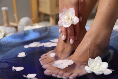 Photo of Woman soaking her feet in bowl with water and flowers on floor, closeup. Spa treatment