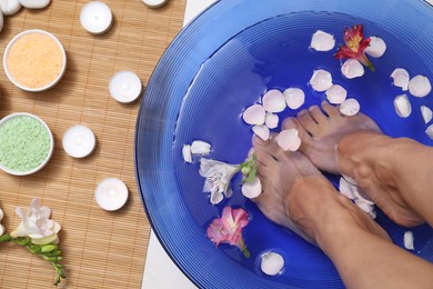 Photo of Woman soaking her feet in bowl with water and flowers on floor, top view. Spa treatment