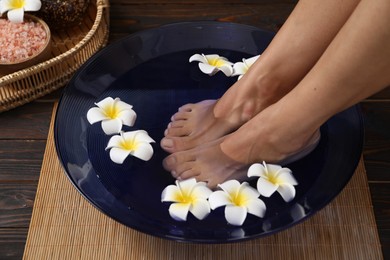 Photo of Woman soaking her feet in bowl with water and plumeria flowers on floor, closeup. Spa treatment