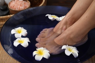 Photo of Woman soaking her feet in bowl with water and plumeria flowers on floor, closeup. Spa treatment