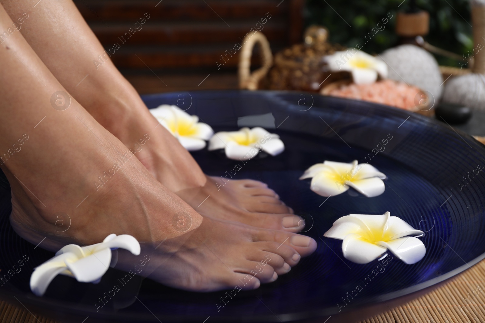 Photo of Woman soaking her feet in bowl with water and plumeria flowers on floor, closeup. Spa treatment