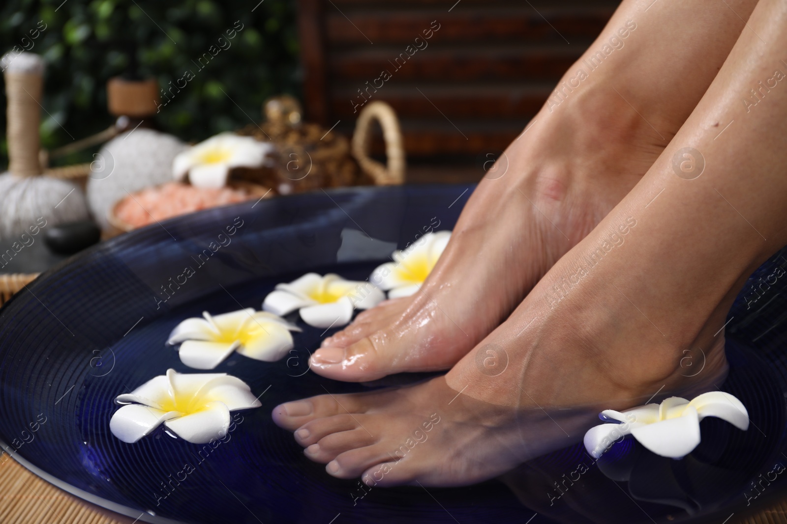 Photo of Woman soaking her feet in bowl with water and plumeria flowers on floor, closeup. Spa treatment