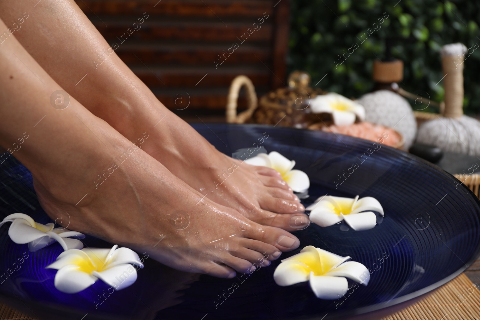 Photo of Woman soaking her feet in bowl with water and plumeria flowers on floor, closeup. Spa treatment