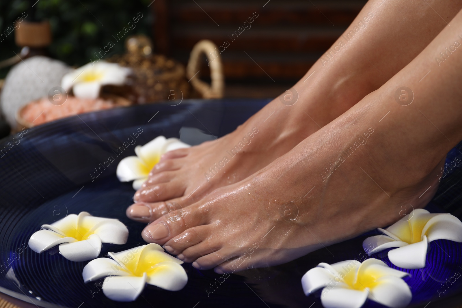 Photo of Woman soaking her feet in bowl with water and plumeria flowers on floor, closeup. Spa treatment