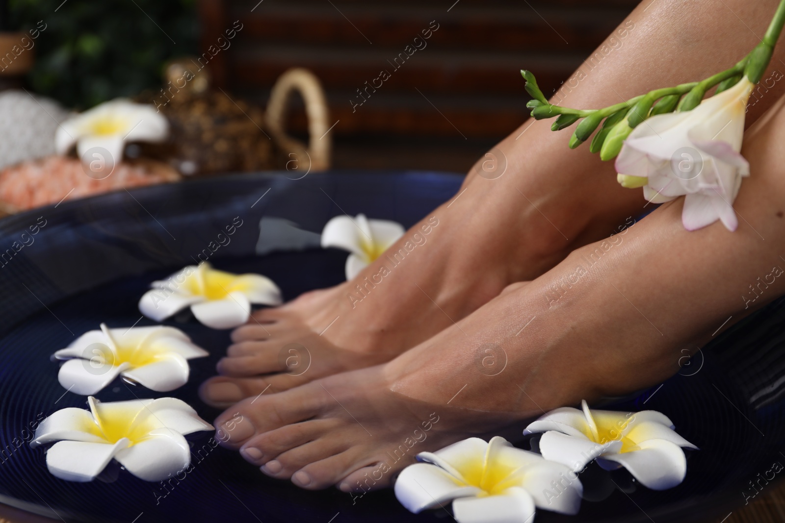 Photo of Woman soaking her feet in bowl with water and plumeria flowers on floor, closeup. Spa treatment